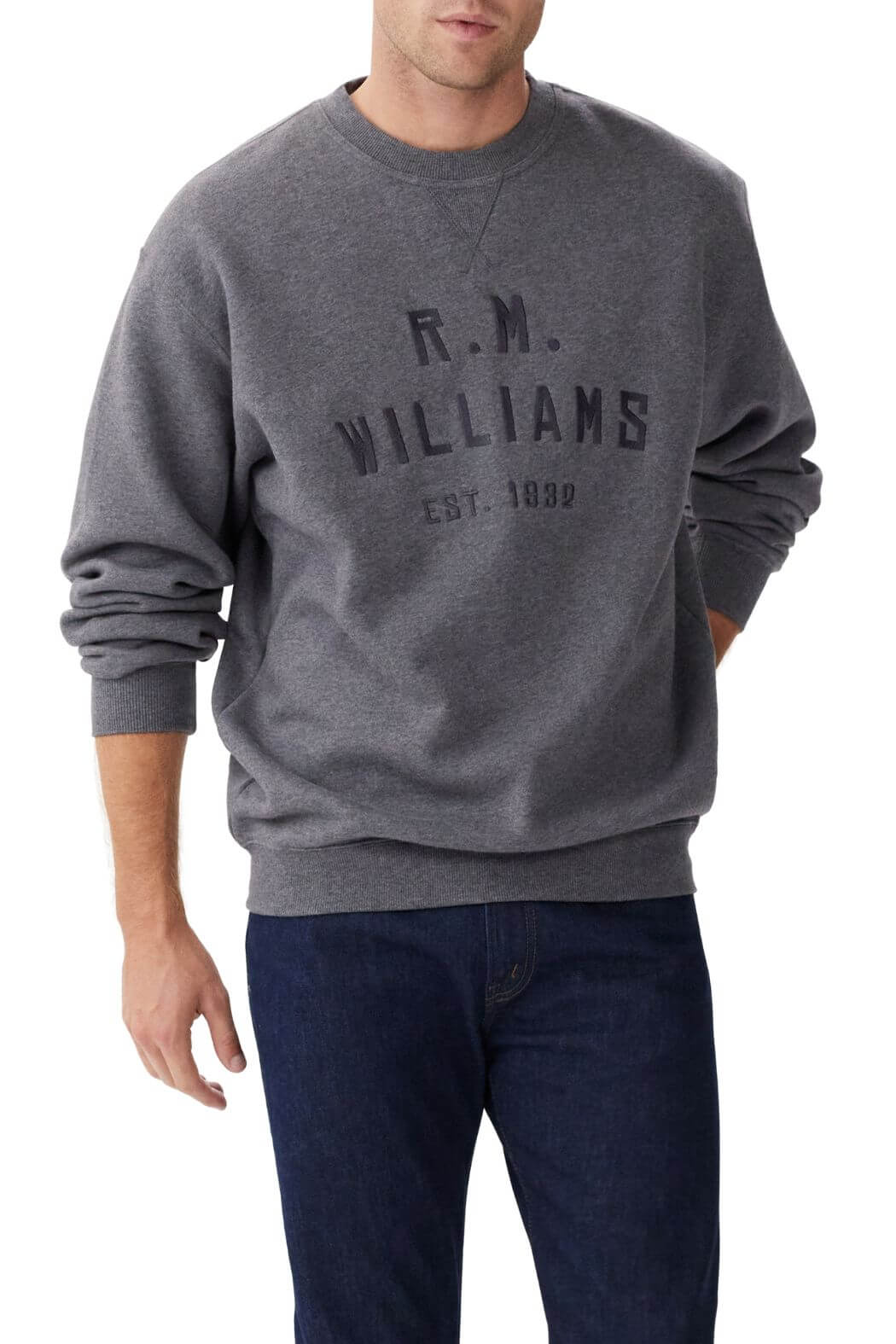 A man wears the RM Williams Bale Sweatshirt, a loose-fitting dark grey fleece with "R.M. Williams Est. 1932" on the front, paired with dark blue jeans—an outback style look against a plain white background.