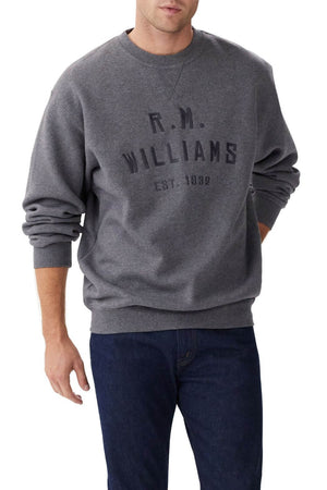 A man wears the RM Williams Bale Sweatshirt, a loose-fitting dark grey fleece with "R.M. Williams Est. 1932" on the front, paired with dark blue jeans—an outback style look against a plain white background.