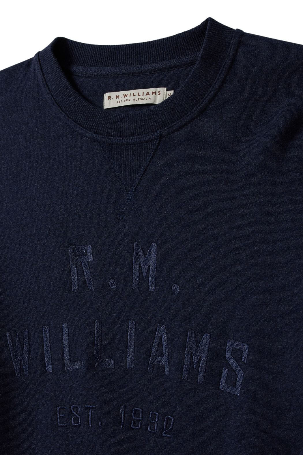 The RM Williams Bale Sweatshirt is a dark blue fleece featuring "R.M. Williams Est. 1932" embroidery on the front, crafted from an organic cotton blend for comfort. The collar label reads "R.M. WILLIAMS," reflecting classic Outback style.