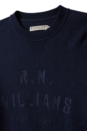 The RM Williams Bale Sweatshirt is a dark blue fleece featuring "R.M. Williams Est. 1932" embroidery on the front, crafted from an organic cotton blend for comfort. The collar label reads "R.M. WILLIAMS," reflecting classic Outback style.