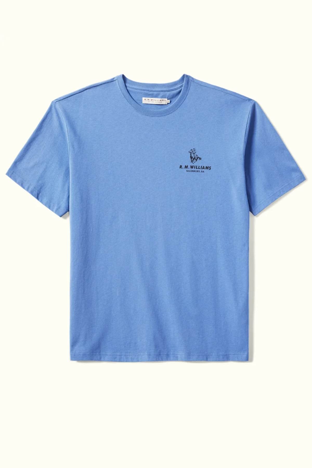 The RM Williams Banner T-Shirt, crafted from organic cotton in a blue hue, showcases a small embroidered design on the left chest with the "R.M. Williams" text and an archive-inspired graphic above it. The shirt is presented flat against a light background.