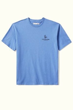 The RM Williams Banner T-Shirt, crafted from organic cotton in a blue hue, showcases a small embroidered design on the left chest with the "R.M. Williams" text and an archive-inspired graphic above it. The shirt is presented flat against a light background.