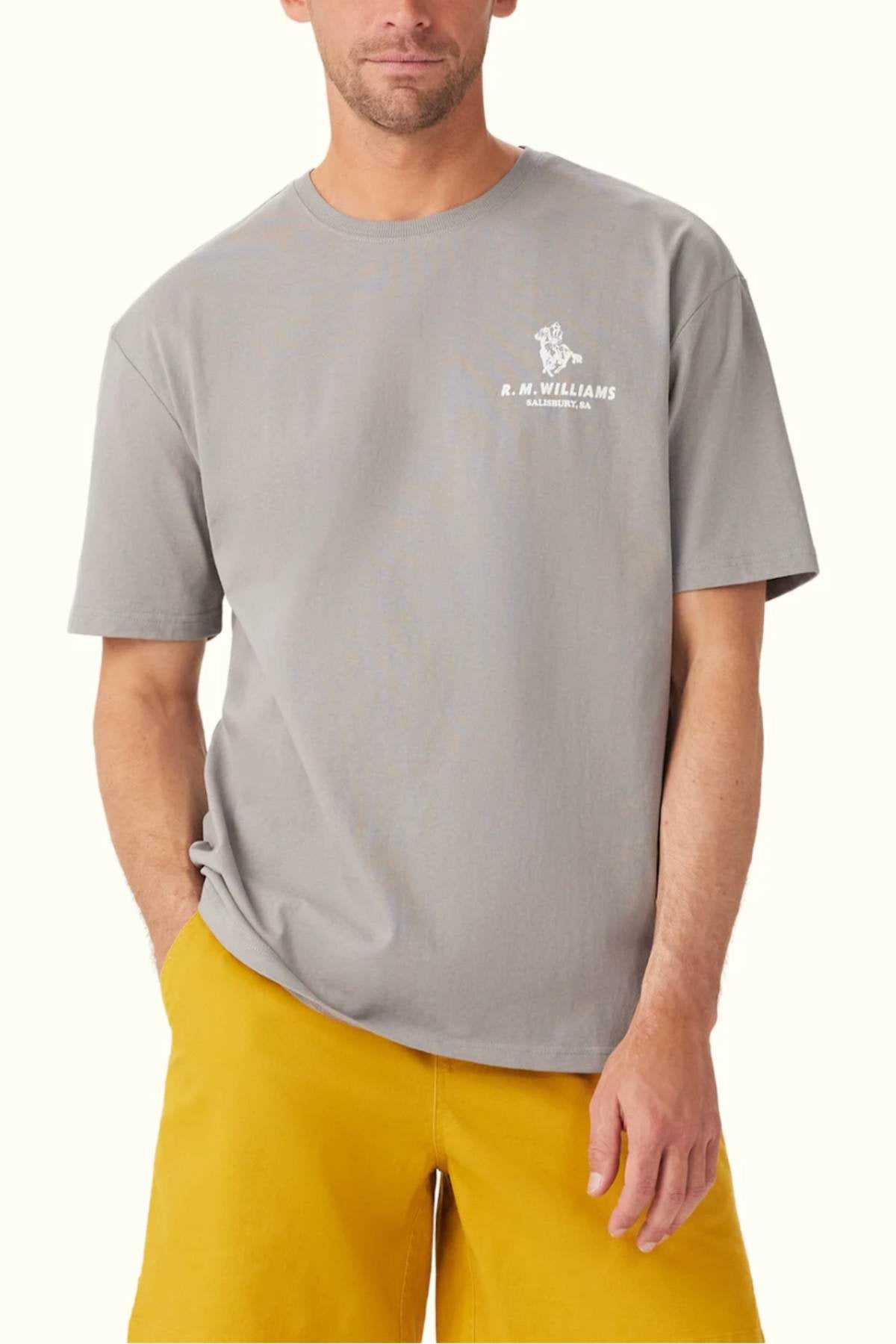 Dressed in a relaxed fit gray RM Williams Banner T-Shirt made from breathable organic cotton and adorned with a subtle heritage logo on the chest, the person pairs it with yellow shorts while standing against a light background, casually placing one hand in their pocket.