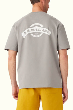 A person wearing a gray RM Williams Banner T-Shirt made from breathable organic cotton, showcasing a heritage logo on the back that says "R.M. Williams, Salisbury Est. 1932, South Australia." This relaxed fit t-shirt pairs perfectly with their yellow pants.