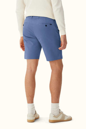 A person is seen wearing a white long-sleeve shirt and the RM Williams Berwick Chino Short, which are modern tailored shorts made from organic cotton. They are paired with white socks and beige sneakers, standing with their back facing the viewer. A plain, light-colored background emphasizes the simplicity and comfort stretch of their outfit.