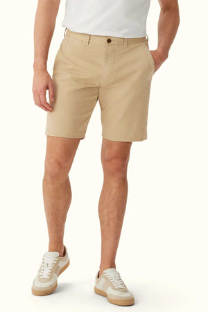 A person wears the RM Williams Berwick Chino Short, elegantly tailored and made from organic cotton, paired with a white shirt and white sneakers adorned with tan details. The shorts feature a button and pockets, offering both style and comfortable stretch as the individual stands against a plain white background.