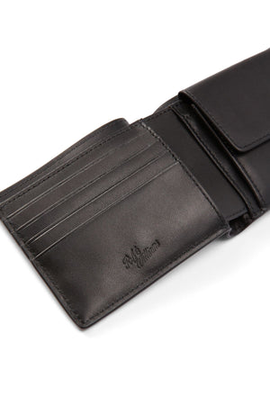 The RM Williams Bi Fold Wallet with Pocket in black full-grain leather is partially open, displaying multiple card slots, a coin pocket, and an embossed “R.M. Williams” logo inside.
