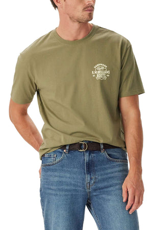 A man wearing the RM Williams Boot T-Shirt in plain olive green, featuring a small white logo on the left chest, paired with blue jeans and standing with one hand in his pocket, looking to the side.