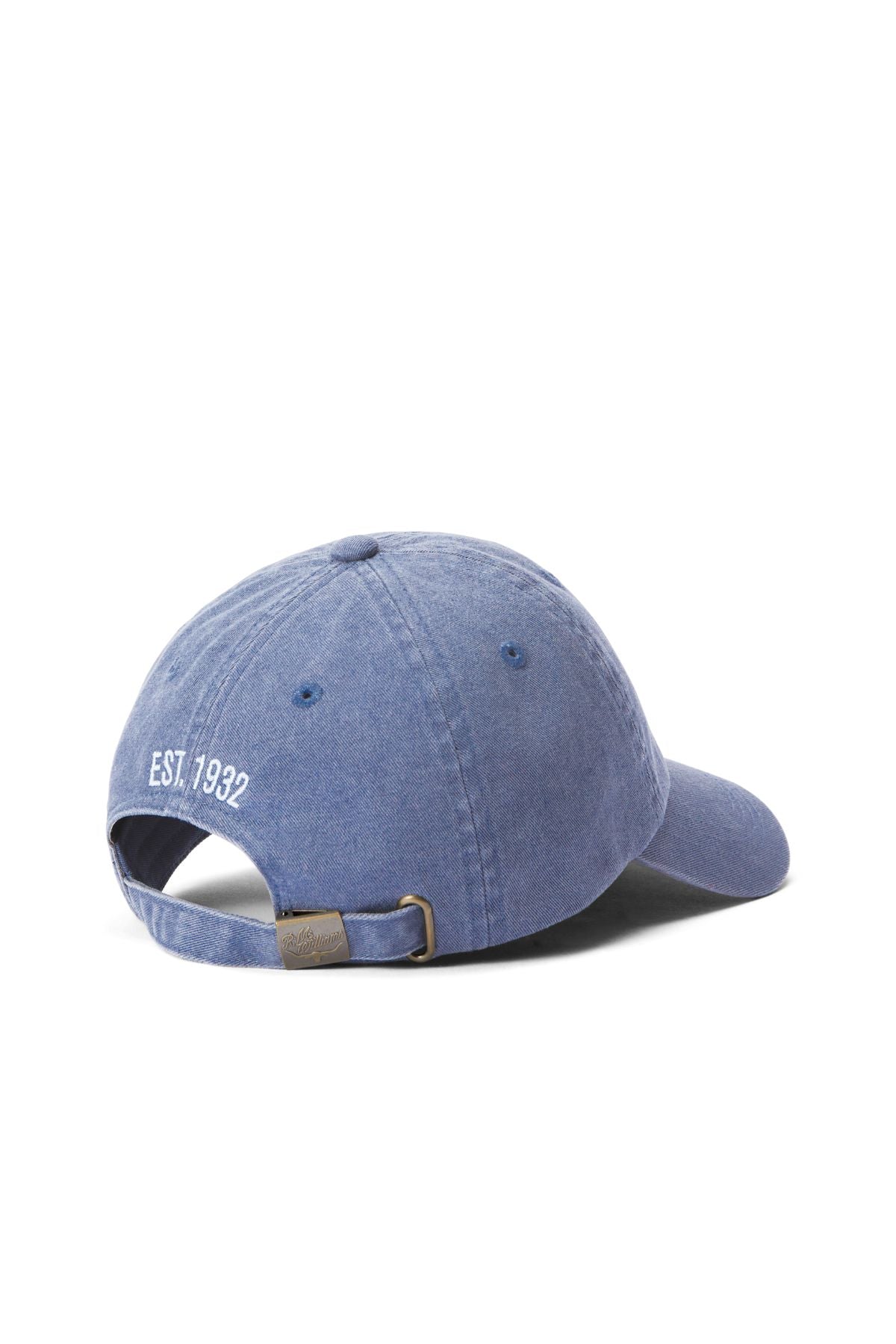 The RM Williams Branded Twill Cap in blue denim organic cotton is shown from the back, displaying a white "EST. 1932" heritage logo above an adjustable strap with a metal buckle.