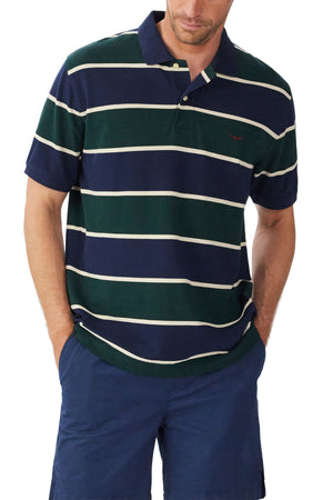A man wears the RM Williams Bright Polo in navy blue and green stripes with white lines and Longhorn embroidery, paired with blue shorts. His hands are in his pockets as he faces slightly downward.