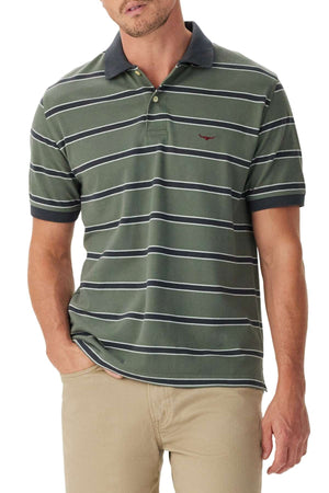 A man wears the RM Williams Brunswick Stripe Polo, featuring green and white stripes with a dark collar and cuffs, paired with beige pants. One hand is in his pocket, and only his upper body is visible.