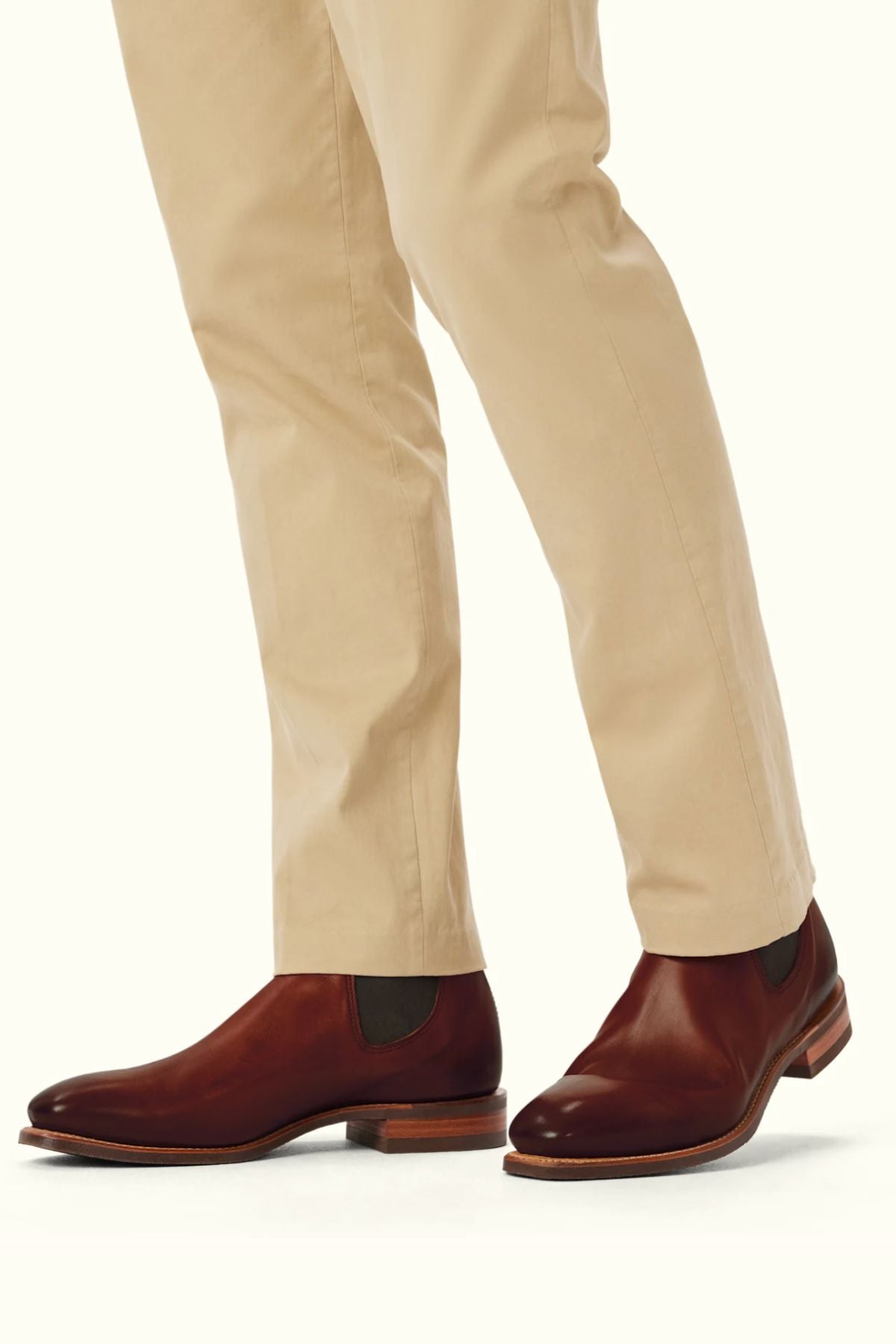 A person in beige pants wears RM Williams Burnished Comfort Craftsman Boots—brown, hand-burnished leather Chelsea boots with a chisel toe—standing on a light background.