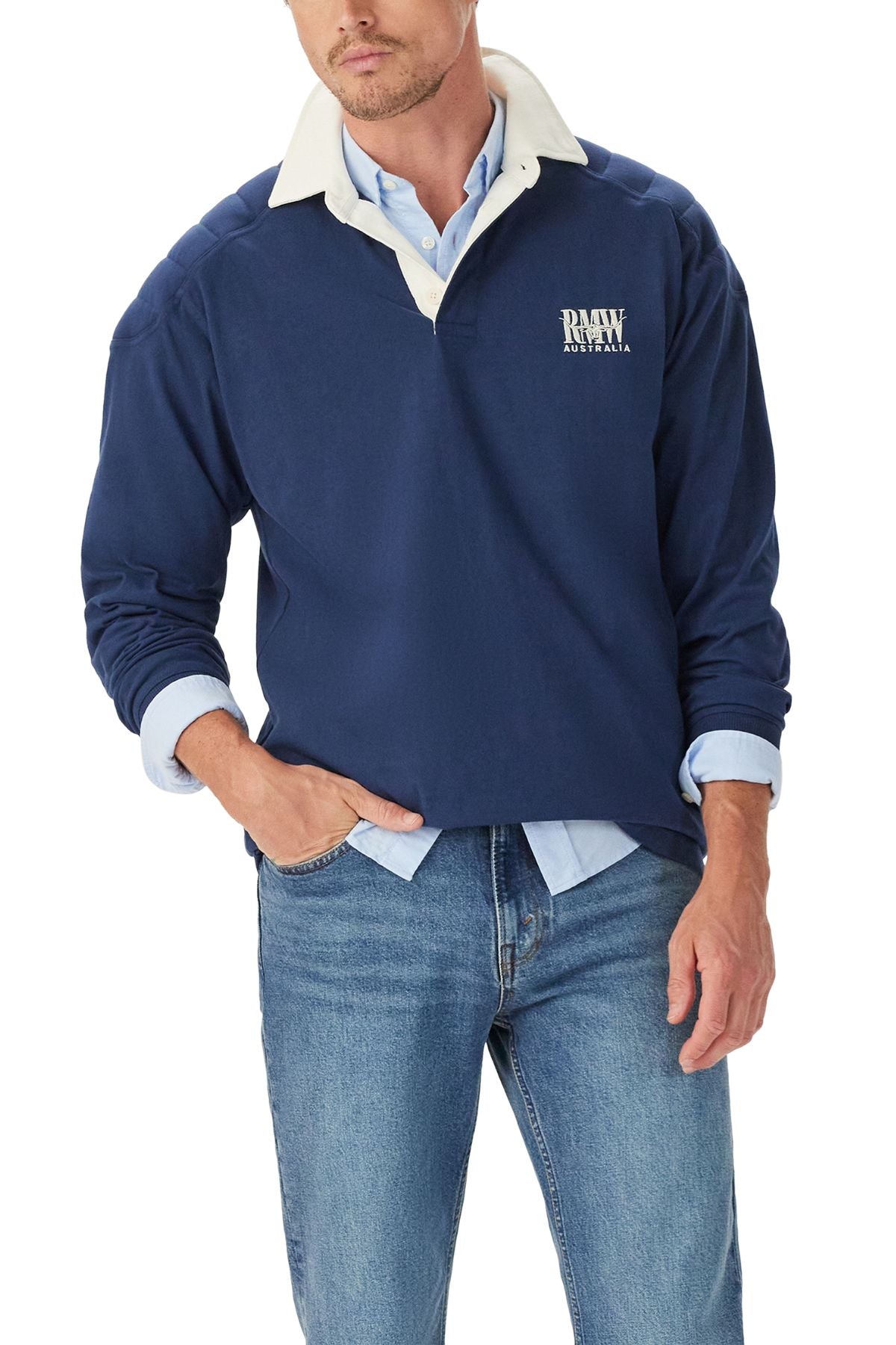 A man wears the RM Williams Classic R.M.W Rugby—navy, with logo embroidery and a white collar—over a light blue shirt and blue jeans, one hand in his pocket.