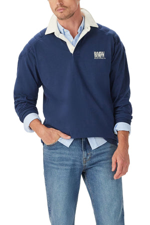 A man wears the RM Williams Classic R.M.W Rugby—navy, with logo embroidery and a white collar—over a light blue shirt and blue jeans, one hand in his pocket.