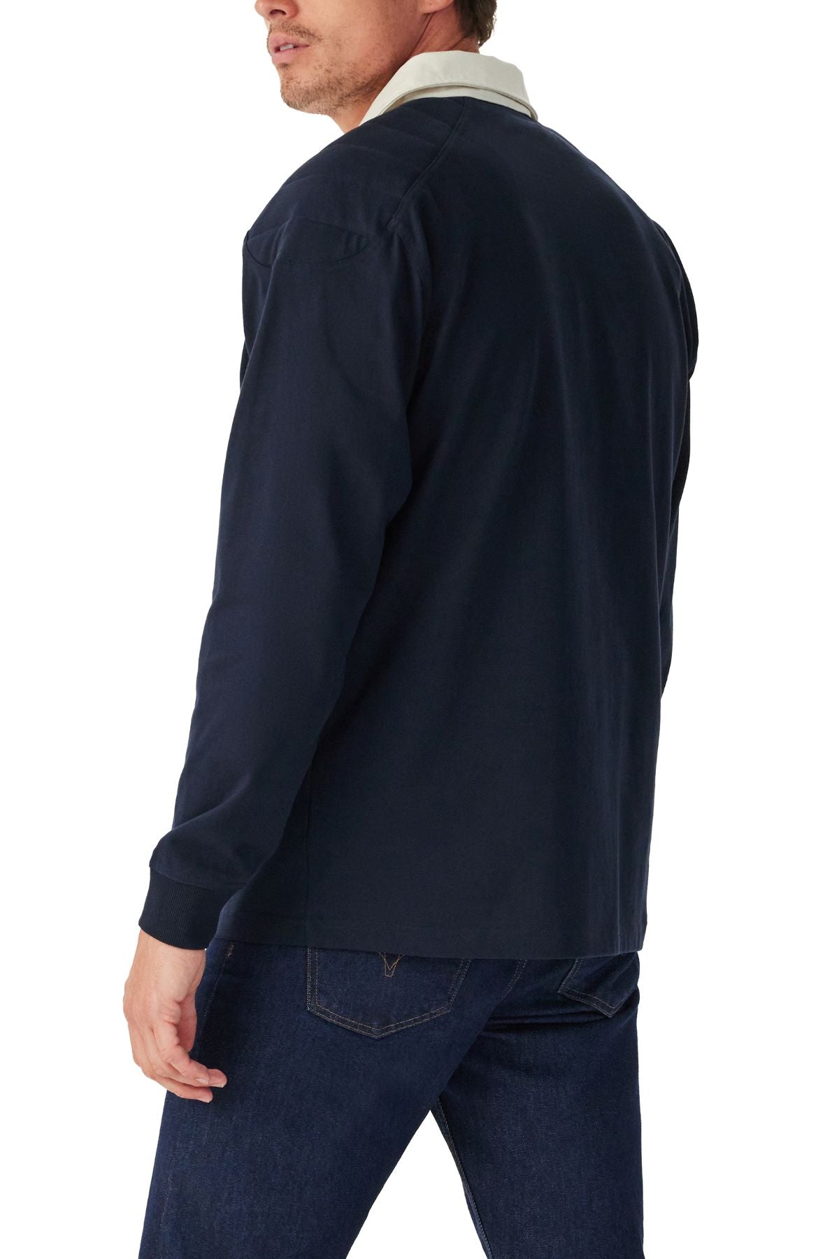 A man in a navy RM Williams Classic R.M.W Rugby, made from heavyweight Australian cotton with a white collar, and dark blue jeans stands facing away from the camera, glancing over his left shoulder.