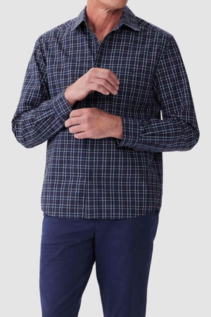A person wearing a RM Williams Classic Shirt, characterized by its dark blue color, long sleeves, checkered pattern, and regular fit, along with dark blue pants is adjusting the cuff of their sleeve. Made from organic cotton, this smart casual attire stands out against the plain, light-colored background. The person's face is not visible.