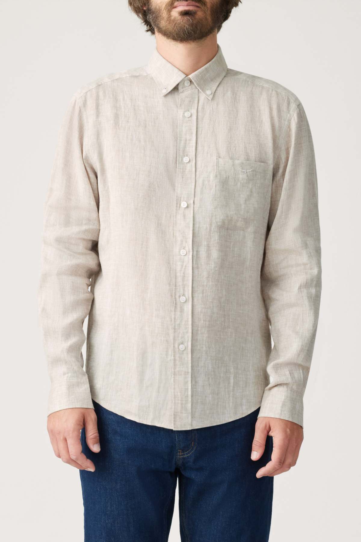 A man with a beard wears an RM Williams Classic Shirt in light beige French linen, featuring a button-down collar and chest pocket, paired with blue jeans. He stands against a plain light background, his face partially out of frame.