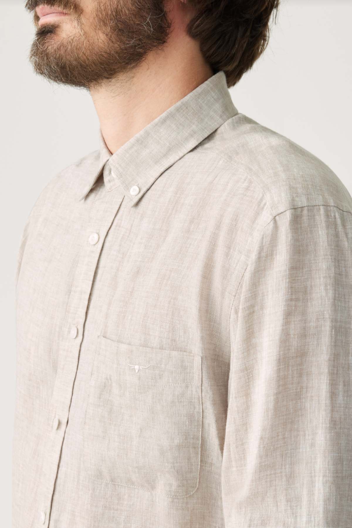 A bearded man faces slightly left against a plain light background, wearing a light beige RM Williams Classic Shirt crafted from French linen, featuring a button-down collar and chest pocket.