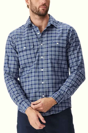 A man wearing a RM Williams Classic Shirt in blue and white plaid stands against a plain light background. He has light facial hair and his hands are clasped in front of him. The smart casual long-sleeved shirt features chest pockets, perfect for a relaxed yet polished look.