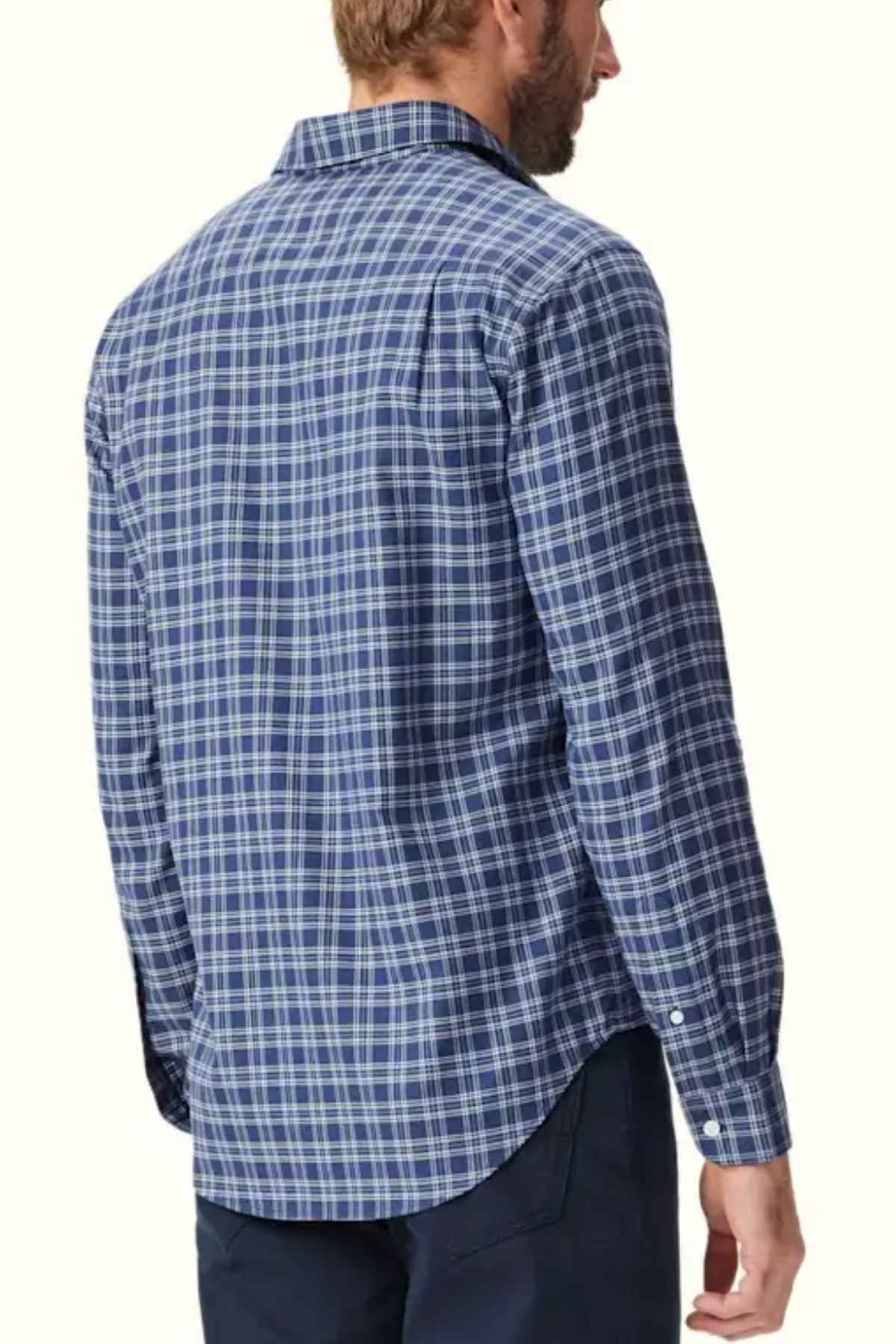 A man with light hair is standing with his back facing the camera, wearing an RM Williams Classic Shirt—a long-sleeved, blue checkered smart casual shirt made of organic cotton and featuring a collar and buttoned cuffs—and dark pants. The background is plain and light-colored.