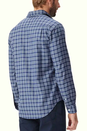 A man with light hair is standing with his back facing the camera, wearing an RM Williams Classic Shirt—a long-sleeved, blue checkered smart casual shirt made of organic cotton and featuring a collar and buttoned cuffs—and dark pants. The background is plain and light-colored.