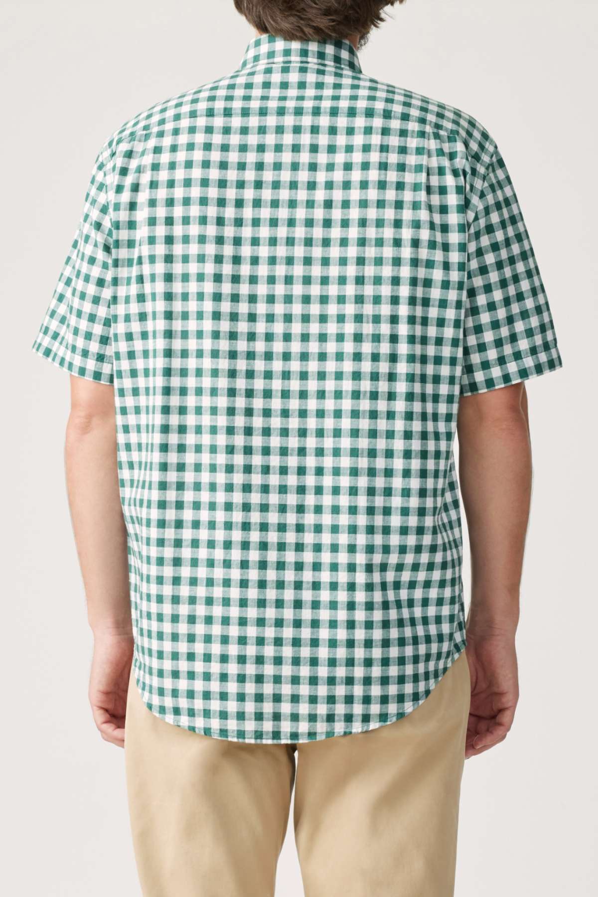A person is wearing an RM Williams Classic Short Sleeve Shirt in green and white gingham, paired with beige pants, viewed from the back. This French linen shirt features a relaxed fit and timeless pattern against a plain white background.