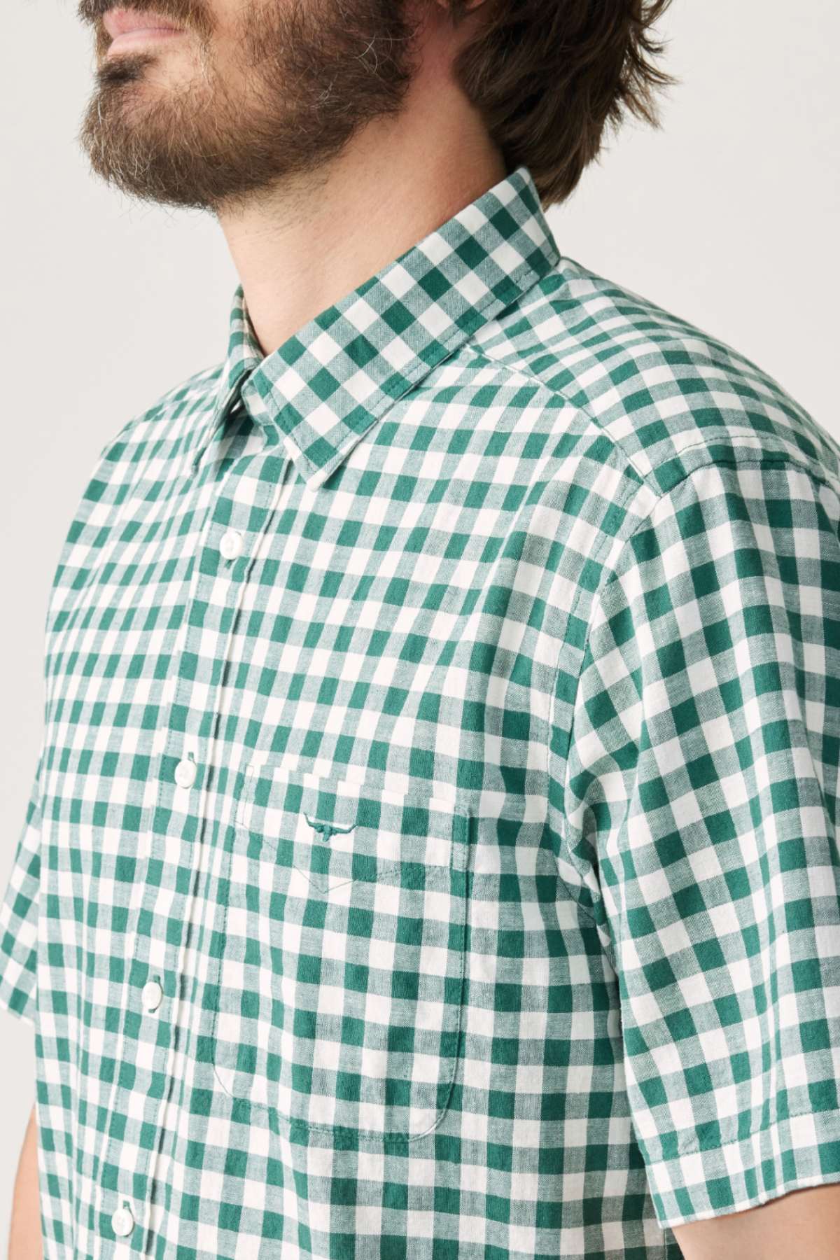 A person wearing the RM Williams Classic Short Sleeve Shirt in a green and white gingham pattern. This French linen shirt includes a pocket on the left side with a small embroidered design. The bearded individual is facing slightly to the left.