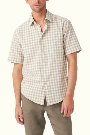 A man dressed in a RM Williams Classic Short Sleeve Shirt, featuring a beige and white gingham pattern, stands against a plain backdrop. This French linen shirt is fully buttoned up, and he pairs it with olive green pants. His head is slightly tilted, displaying a neutral expression.