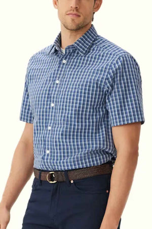 A man wearing an RM Williams Classic Short Sleeve Shirt, featuring a blue and white checkered pattern, regular fit, and classic collar with white buttons. The shirt is tucked into dark blue pants secured with a brown belt. The background is plain and light-colored.