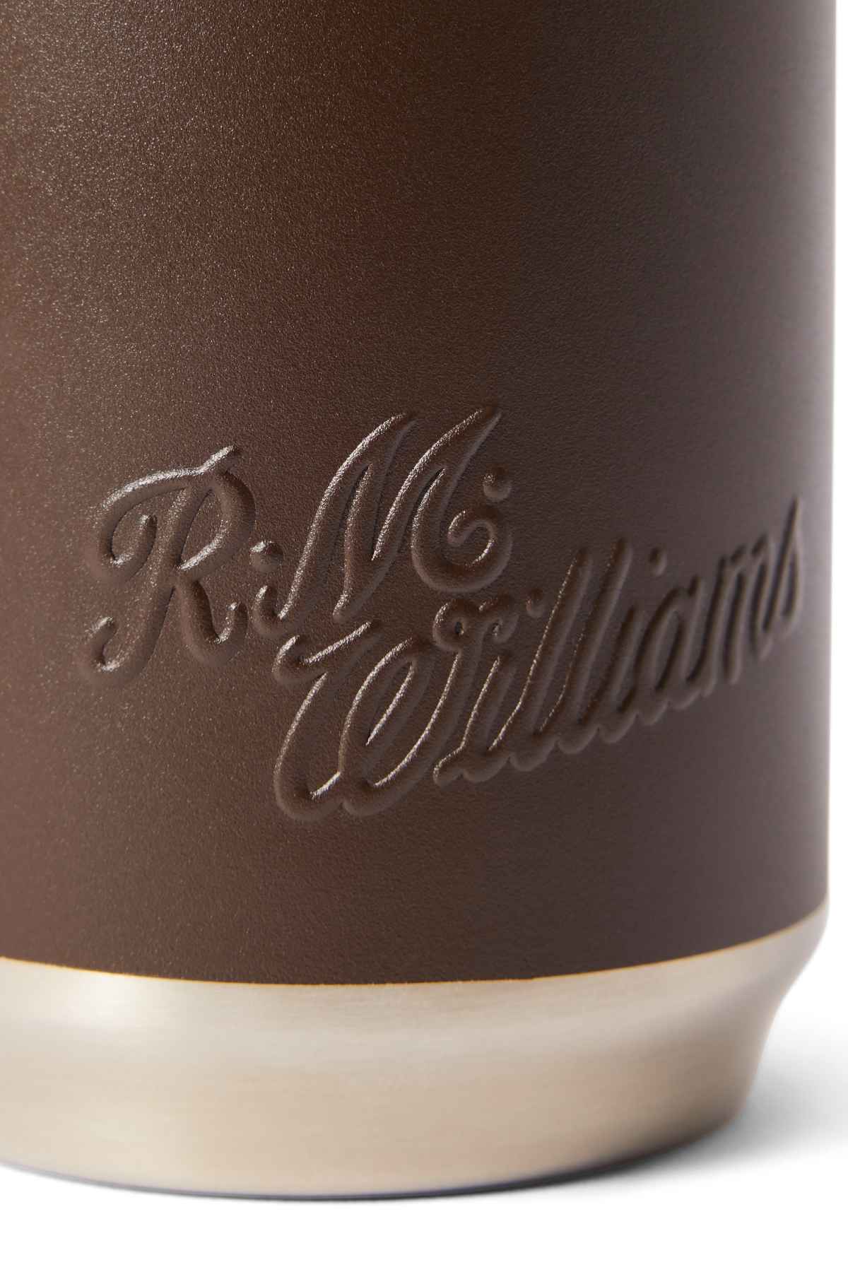Close-up of a textured brown leather surface embossed with "R.M. Williams" in cursive, akin to their RM Williams Coffee Cup. The base is light-colored, metallic or plastic, suggesting sustainability.