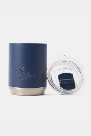 The RM Williams Coffee Cup, featuring a navy blue insulated body, stainless steel base, and embossed logo, is displayed beside its clear plastic lid with sliding closure on a plain white background.