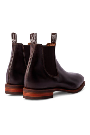 A pair of RM Williams Comfort Craftsman boots in dark brown leather with a polished finish. These Chelsea boots feature elastic side panels and pull tabs with white text at the back. Made in Australia, they boast wooden soles with a layered design. The image shows a rear view of the boots.