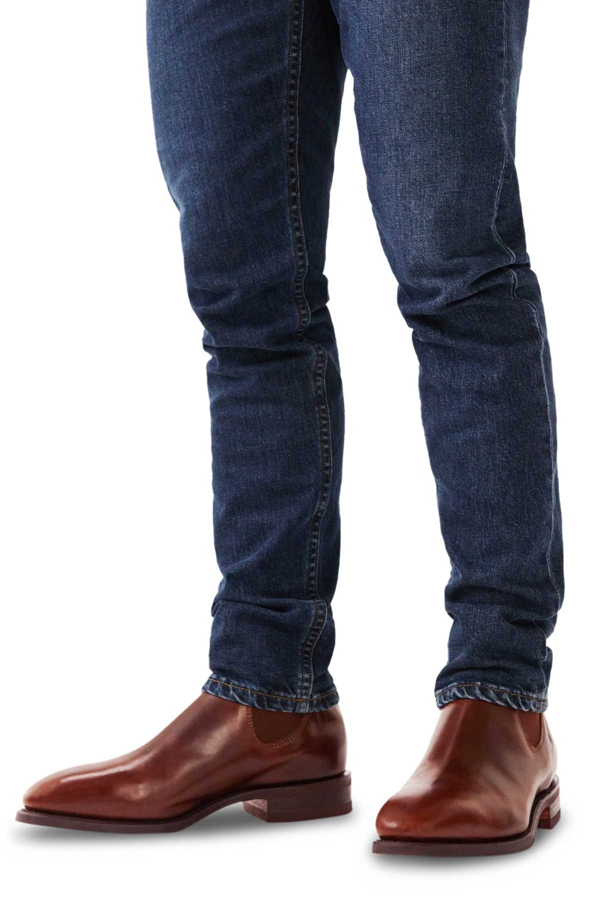 A person wearing dark blue jeans and RM Williams Comfort Craftsman Boots in brown leather with a rubber welt sole, standing against a white background.