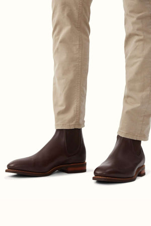A person wearing light beige, slightly wrinkled pants and dark brown RM Williams Comfort Kangaroo Craftsman Boots made in Australia with a polished finish. The boots, crafted from kangaroo leather, feature elastic side panels and a light brown sole with a slight heel. The background is plain white.