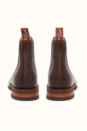 A pair of RM Williams Comfort Kangaroo Craftsman Boots seen from the back, crafted from premium kangaroo leather. Each boot includes a tab with white text at the top and showcases a wooden outsole with a reddish-brown hue, complemented by a rubber heel for enhanced grip. They are positioned side by side against a plain, light background.