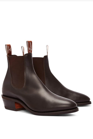 The RM Williams Comfort Lady Yearling Rubber Sole Boot is a pair of dark brown leather Chelsea boots with elastic side panels and pull tabs, featuring a block heel and a smooth finish.