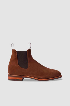 The RM Williams Comfort Suede Craftsman Boot features brown suede leather, black elastic side panels, a low wooden heel, and front and back pull tabs, showcased against a plain light gray background.
