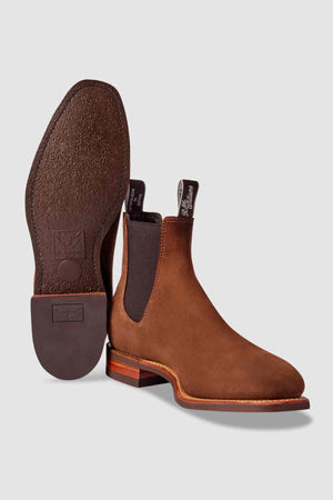 The RM Williams Comfort Suede Craftsman Boot is a pair of brown suede Chelsea boots with elastic side panels, pull tabs, textured sole, and wooden heel. One boot stands upright, the other on its side; classic Australian craftsmanship.