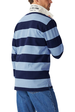 A man is seen from the back, turned slightly to the side, wearing blue jeans and the RM Williams Cradle Rugby—a long-sleeve organic heavyweight rugby shirt with blue and navy stripes, white collar.