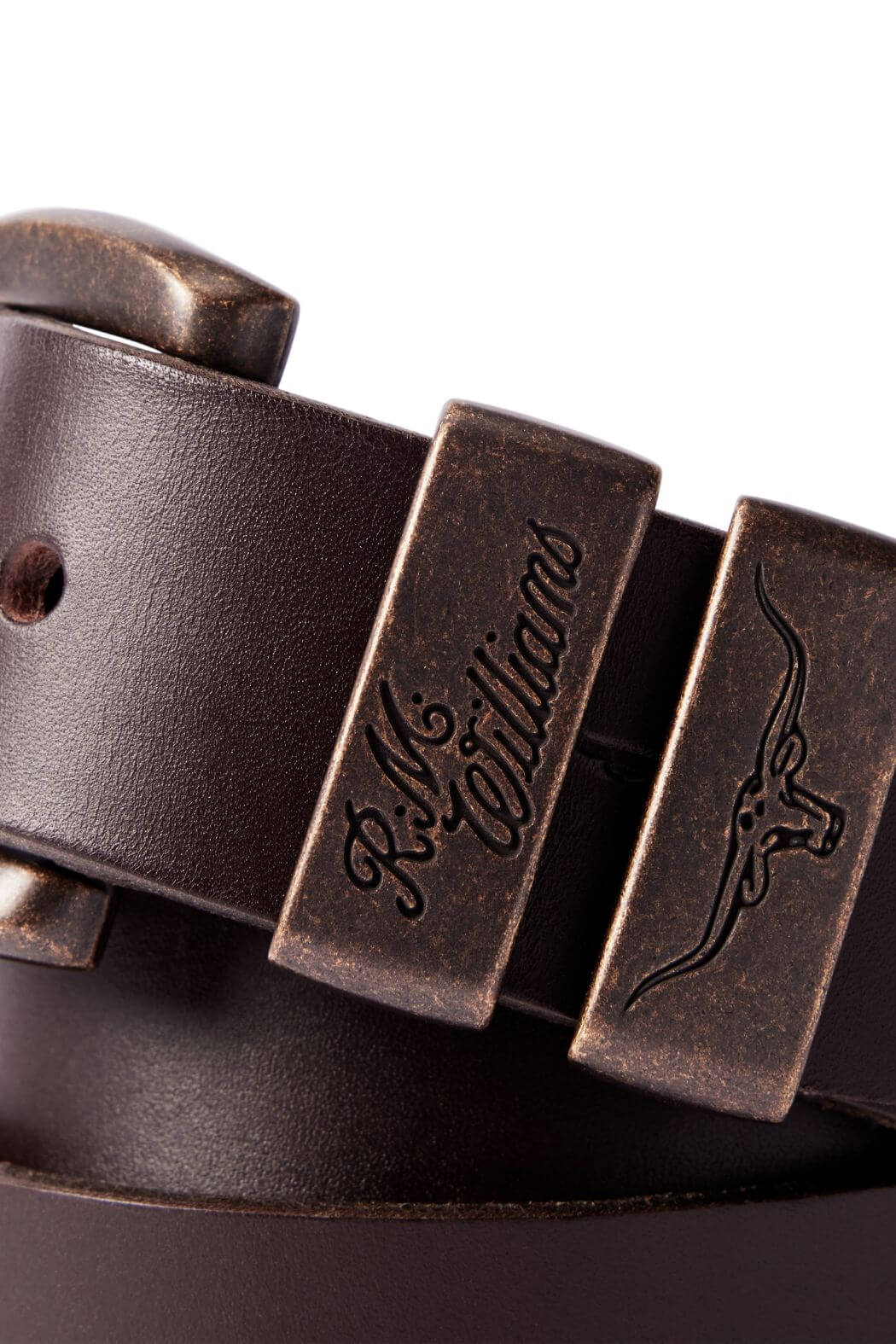 Close-up of the RM Williams Drover Belt in brown, featuring an antique brass buckle adorned with engraved details such as "R.M. Williams" text and a longhorn steer image. The vegetable-tanned leather exhibits a smooth finish, with an adjustable hole visible.