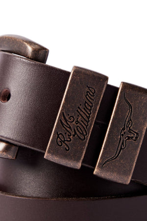 Close-up of the RM Williams Drover Belt in brown, featuring an antique brass buckle adorned with engraved details such as "R.M. Williams" text and a longhorn steer image. The vegetable-tanned leather exhibits a smooth finish, with an adjustable hole visible.