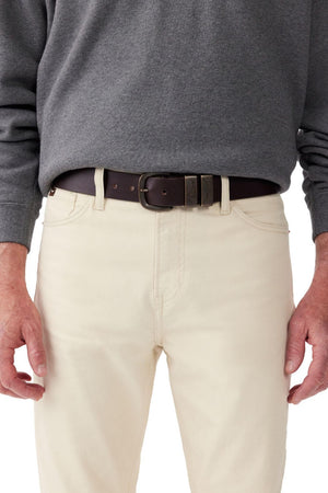 A person wearing a gray long-sleeved shirt and light beige pants is shown in this image from the waist down. They sport a dark brown, vegetable-tanned leather RM Williams Drover Belt with an antique brass buckle. Their hands are relaxed at their sides.