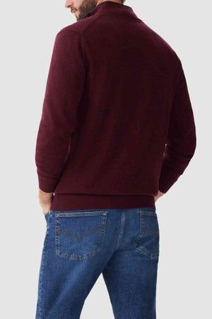 A man is standing with his back to the camera, wearing a maroon RM Williams Ernest Sweater and blue jeans. His left hand is in his pocket, and the background is a light grey color.