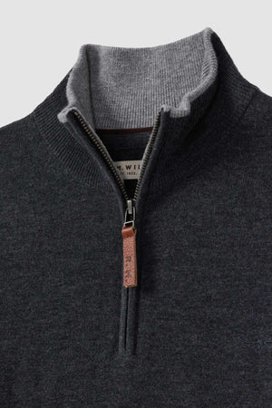 A close-up of an RM Williams Ernest Sweater in dark gray shows off the fine gauge merino quarter-zip design, complemented by a lighter gray inner collar. The zipper is accented with a leather pull tab. A small portion of the inner collar label is visible, displaying "WIL" and "192T." The sweater looks to be made of a soft material.