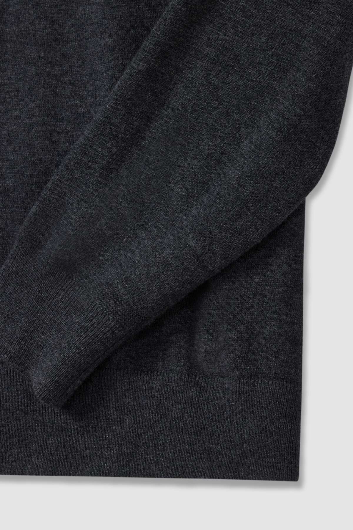 Close-up image of a dark gray RM Williams Ernest Sweater sleeve and partial torso, showcasing the texture and knit pattern of the fine gauge merino fabric. The neutral background highlights the soft and cozy material of this garment.