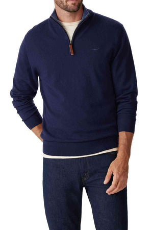 A man wears the RM Williams Ernest Sweater in navy blue merino wool over a white shirt and dark blue jeans, standing with one hand in his pocket against a plain white background.