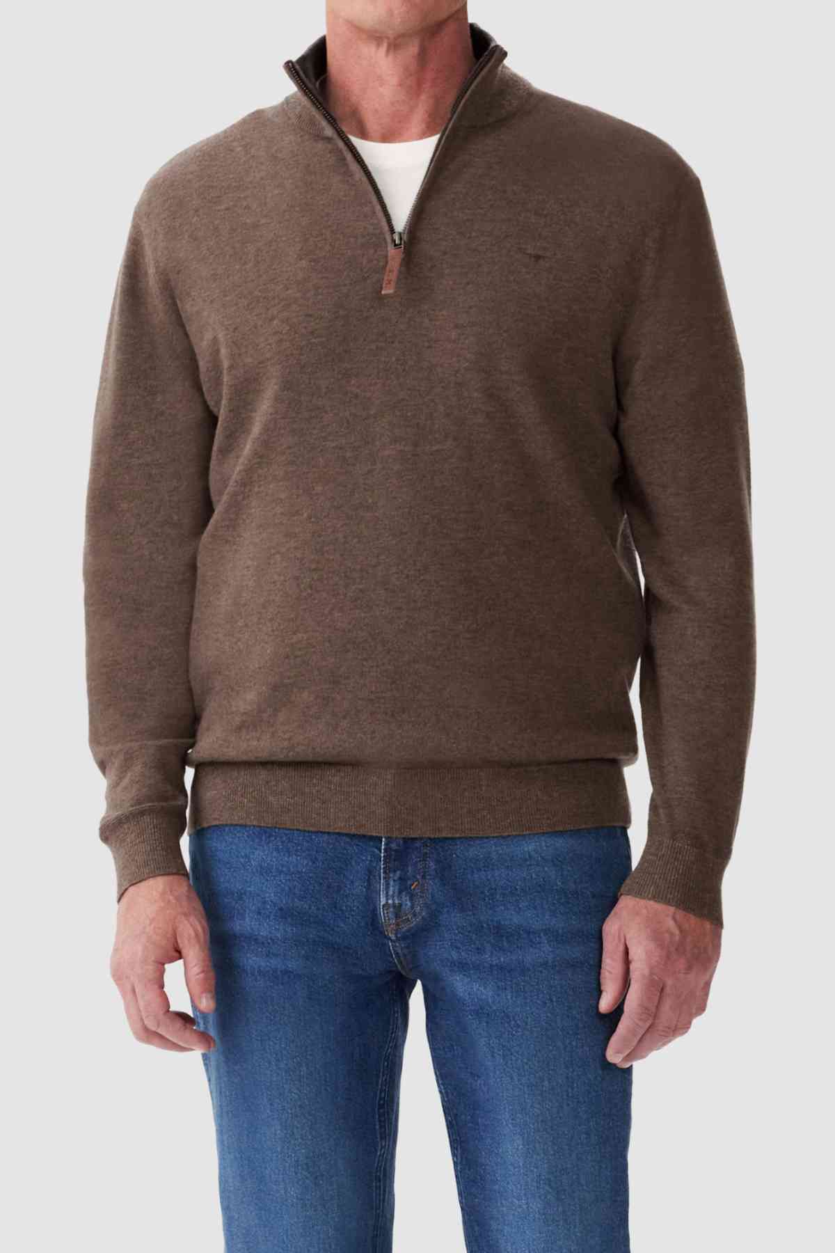 A person in an RM Williams Ernest Sweater stands against a plain background, wearing a fine gauge brown merino quarter-zip sweater over a white shirt and blue jeans. The luxurious Merino wool sweater has a slightly raised collar with the zipper partially open.