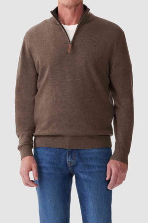 A person in an RM Williams Ernest Sweater stands against a plain background, wearing a fine gauge brown merino quarter-zip sweater over a white shirt and blue jeans. The luxurious Merino wool sweater has a slightly raised collar with the zipper partially open.