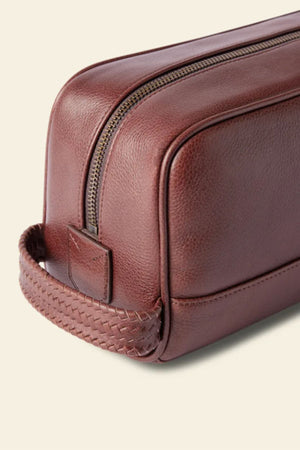 A close-up of the RM Williams Farrier Signature Washbag in brown tumbled grain leather with a woven handle and gold zipper, set against a light beige background.