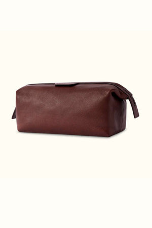 The RM Williams Farrier Washbag is a rectangular brown leather washbag with a top zipper closure. Crafted from tumbled grain leather, it showcases a simple, smooth design and is elegantly set against a light neutral background.